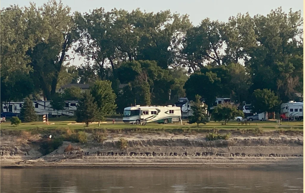A green and white motorhome sits lit up by the sun along a river bank
