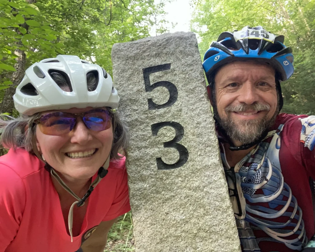 About A couple dressed in bike gear and bike helmets smile beside a 53 mile marker sign