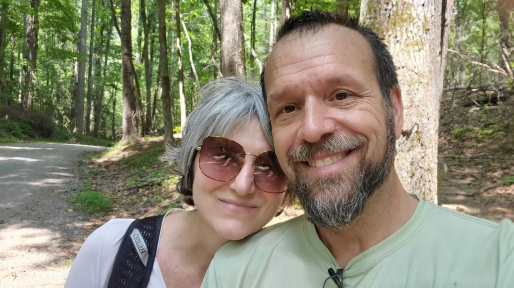 About A couple stands in the woods smiling out on a trail.