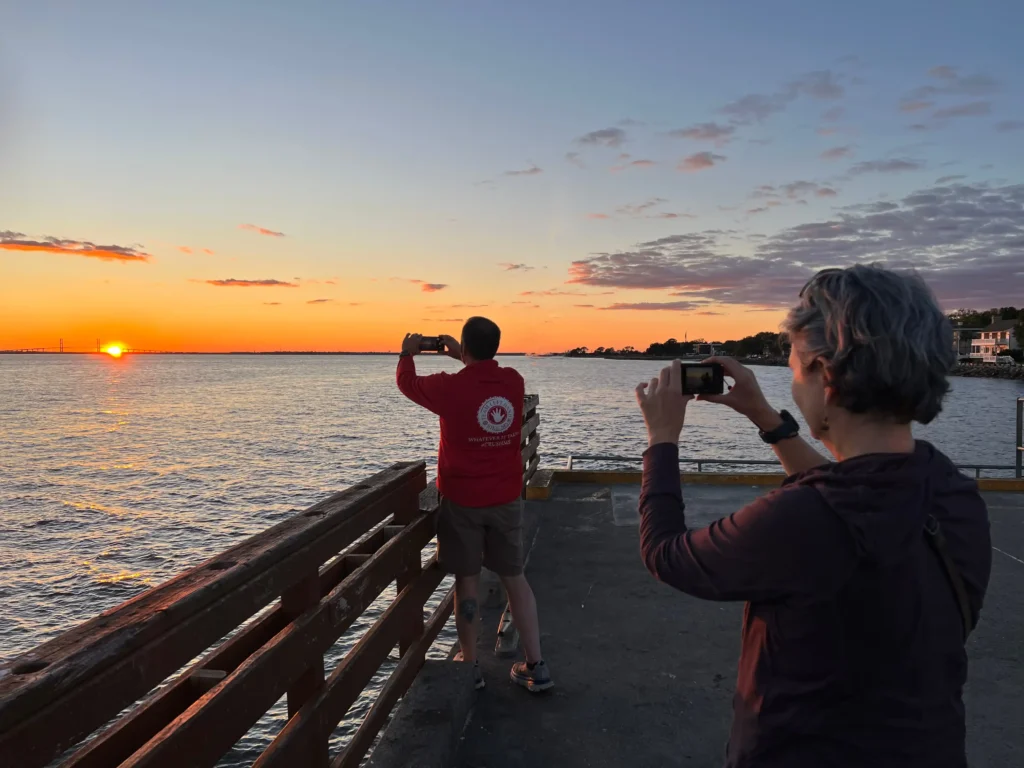 About An couple stands on a pier at sunset, each holding a camera as they photograph the colorful sky.