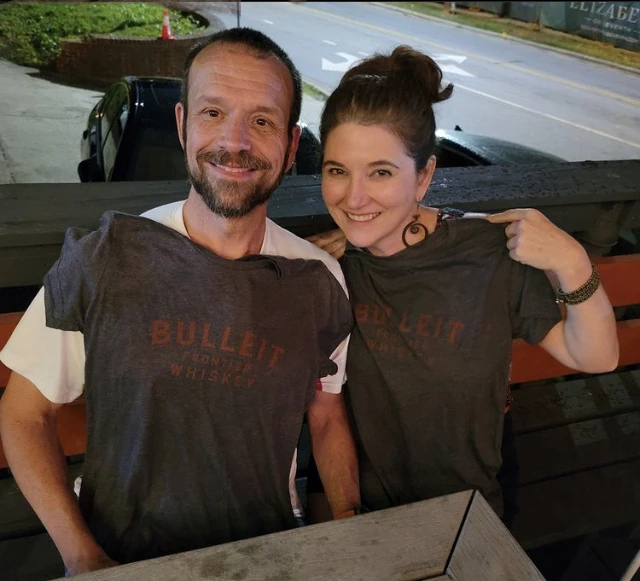 About A couple is seated and smiling while showing off matching t-shirts