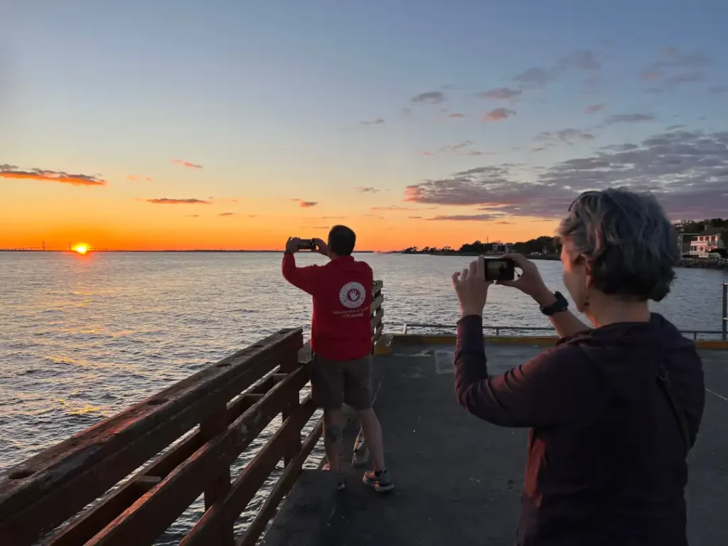 A couple stands on a pier at sunset, each holding a camera as they photograph the colorful sky.
