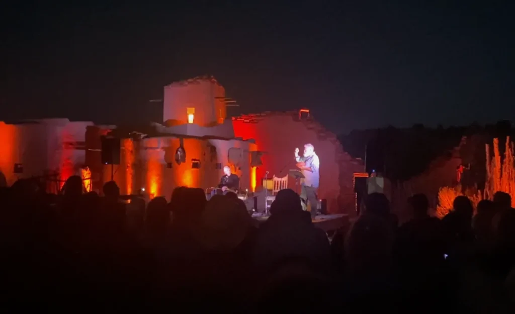Two performers, one standing and one seated perform on a stage in front of a dramatic red lit adobe building.