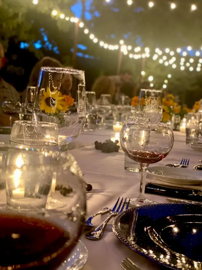 Wine glasses shimmer on a long table lit in the evening by strands of lights