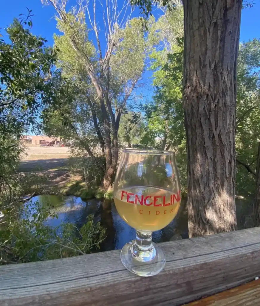 A glass of cider sits on a railing with a view of a river behind it.